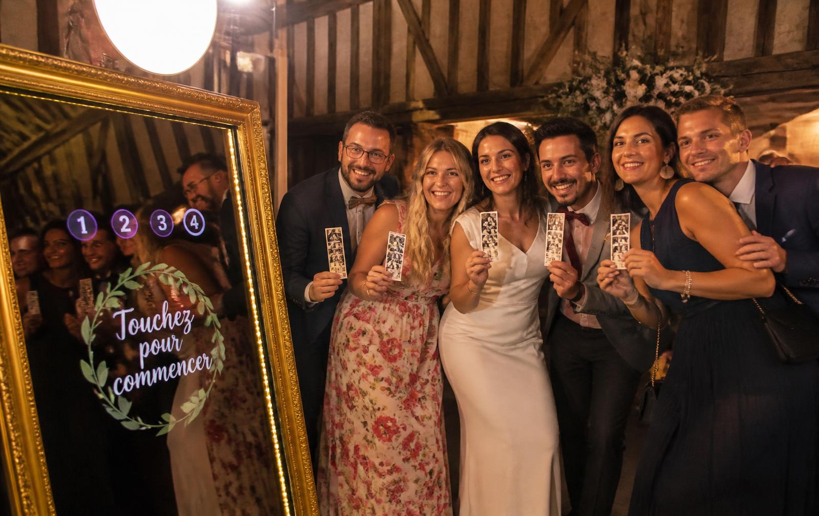 Invités d’un mariage normand souriant avec leurs bandes photo devant un photomaton miroir doré dans une grange à colombages